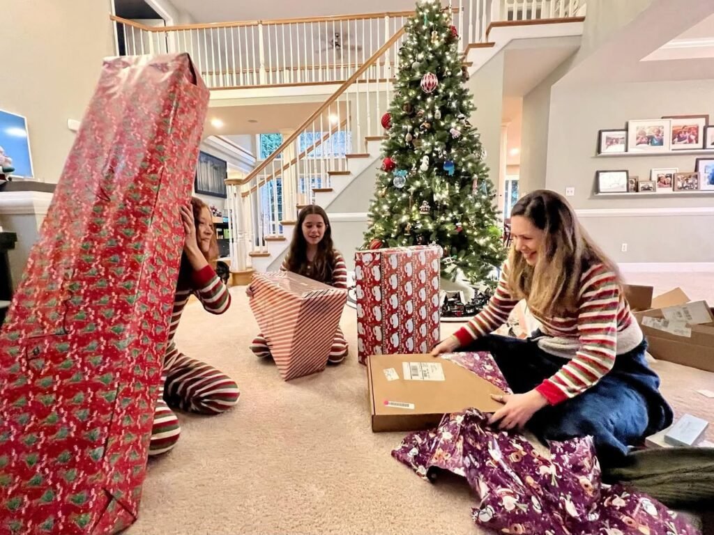 A family wraps gifts together on the floor near a decorated Christmas tree with wrapping paper and boxes.