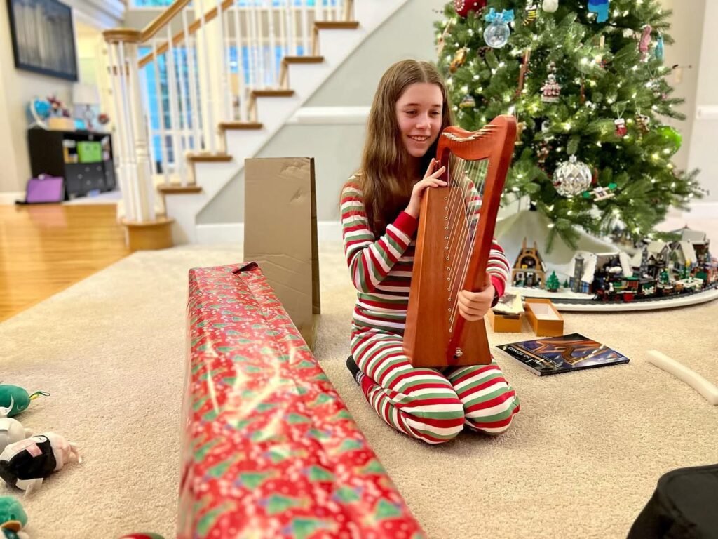 A person kneeling on the floor playing a small wooden harp next to a decorated Christmas tree.