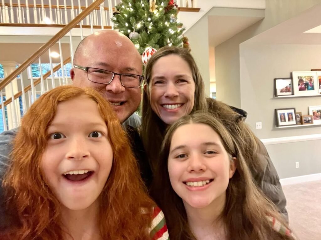 A family smiles joyfully for a holiday photo in front of a decorated tree and staircase. Festive photos are visible on the walls in the background.