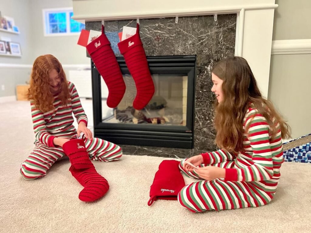 Two children in striped pajamas sit by a fireplace with red Christmas stockings.
