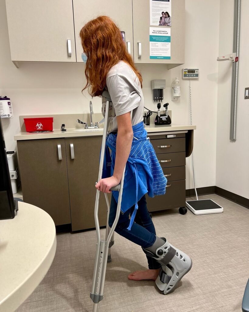 A person with red hair walks through a hospital hallway using crutches and wearing a walking boot.