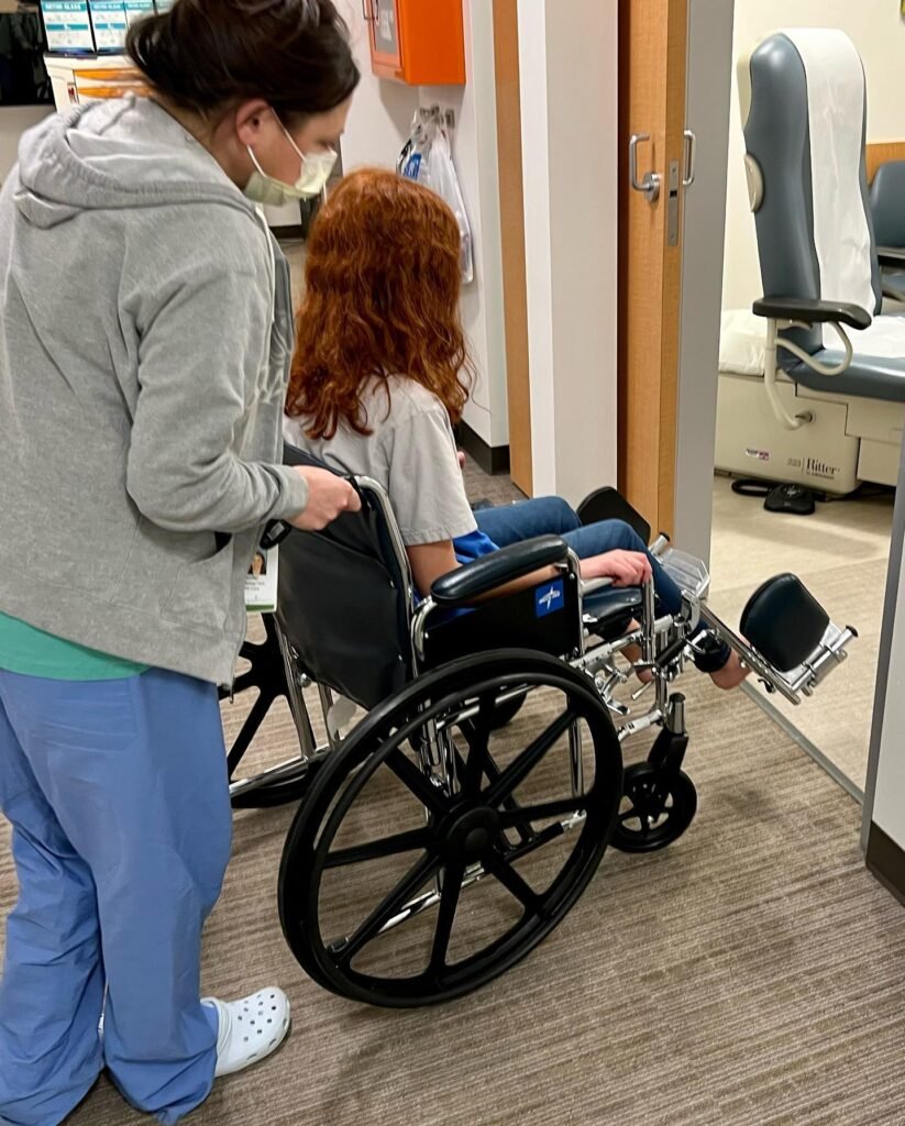 A caregiver in scrubs and a mask accompanies a person in a wheelchair in a hospital room.