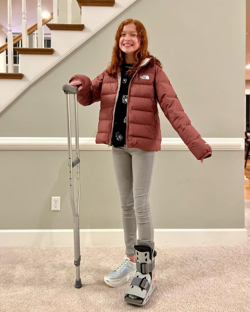 A person in a puffer jacket and walking boot stands smiling indoors while holding a crutch. Behind them is a carpeted room and a staircase.