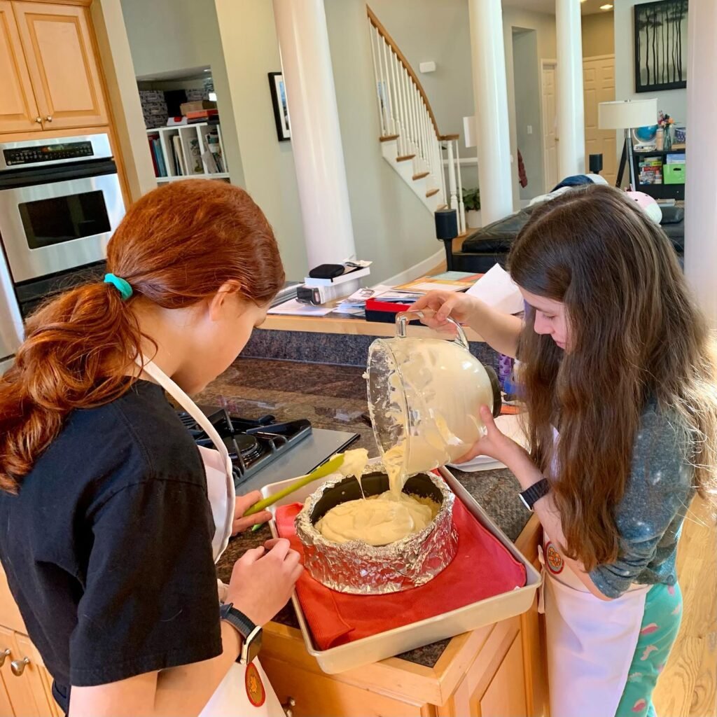 Children pouring cake batter into a pan covered in foil on a red-lined tray in a modern kitchen.