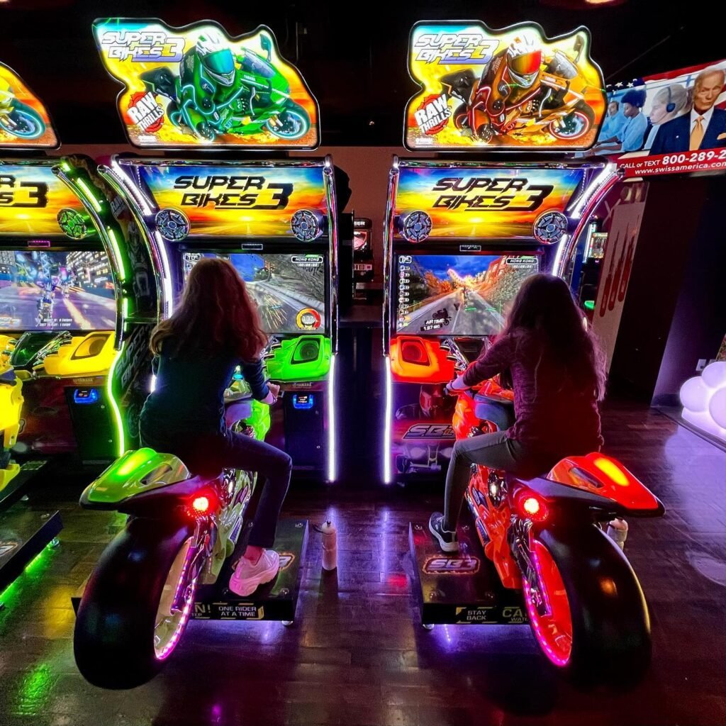 Two people are riding illuminated motorcycle arcade games at an arcade venue. The Super Bikes 3 cabinets feature colorful neon lights and screens.
