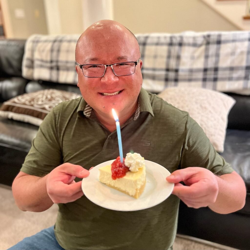 A smiling person holds a plate with a slice of lemon cake and a lit blue candle. The background includes a cozy living room with a plaid blanket on the couch.