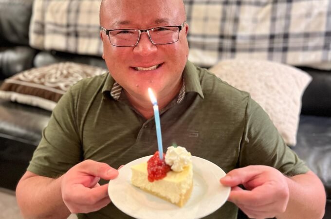 A smiling person holds a plate with a slice of lemon cake and a lit blue candle. The background includes a cozy living room with a plaid blanket on the couch.