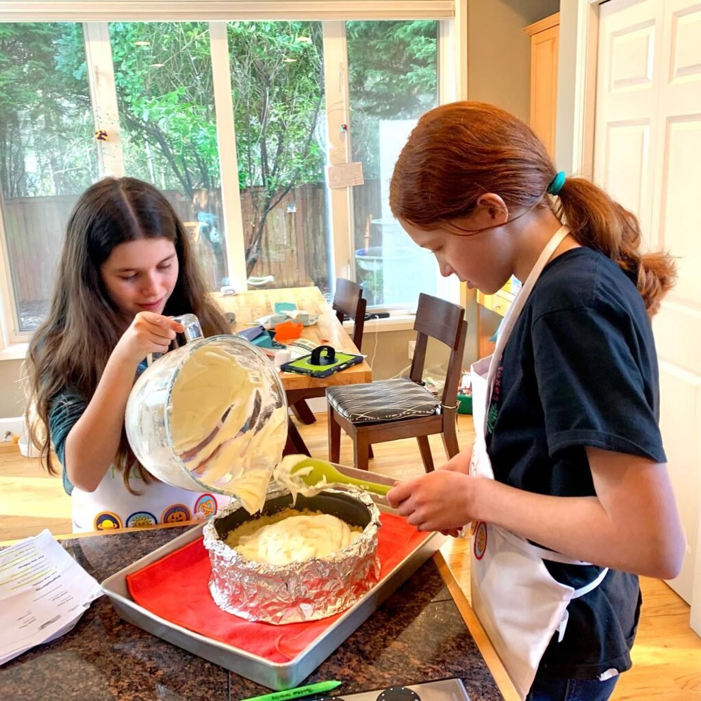 Two children wearing aprons prepare a cake in a bright, natural-lit kitchen while one pours batter into a bundt pan.