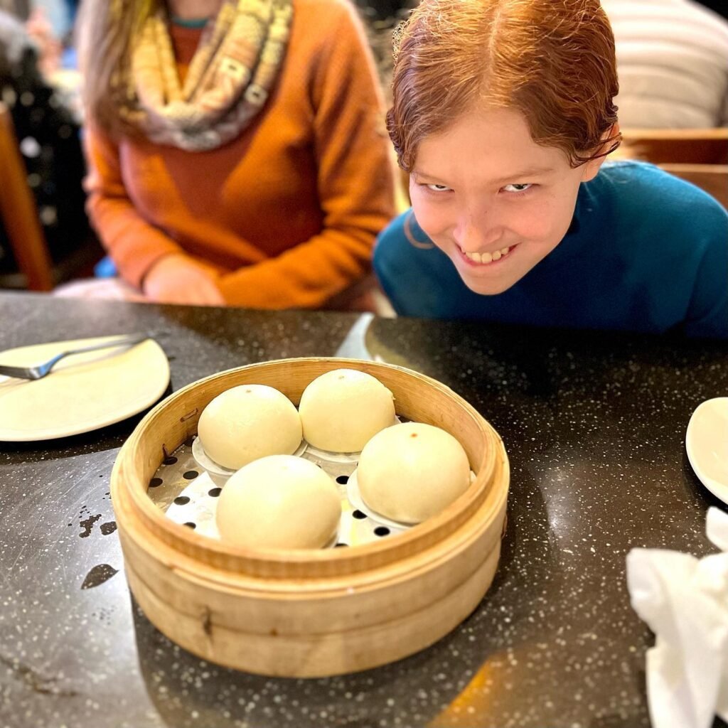 A wooden steamer basket filled with steamed dumplings on a table. A smiling person in a blue shirt sits next to the steamer in a restaurant setting.