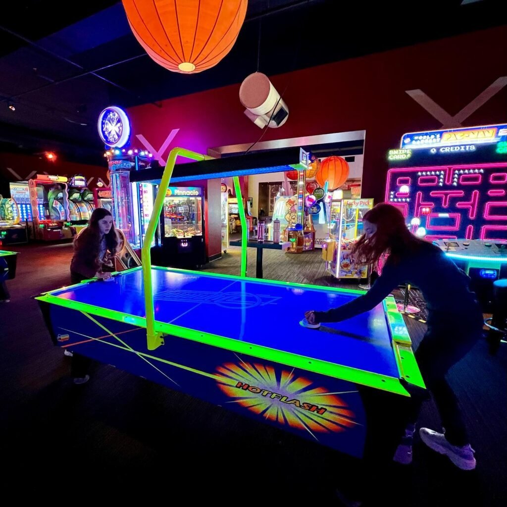 Two players compete in a neon-lit air hockey game under colorful pendant lights and arcade machines.