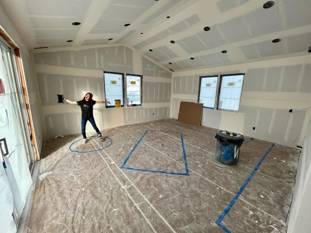A person stands in an unfinished room with freshly installed drywall, underlayment floor, and a coffered ceiling. Blue tape outlines sections on the floor.