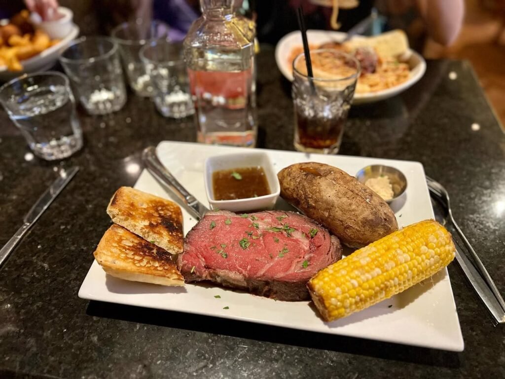 A delicious served plate of steak, corn, and potatoes on a dark dining table. Accompanied by water glasses and condiments for a satisfying meal.