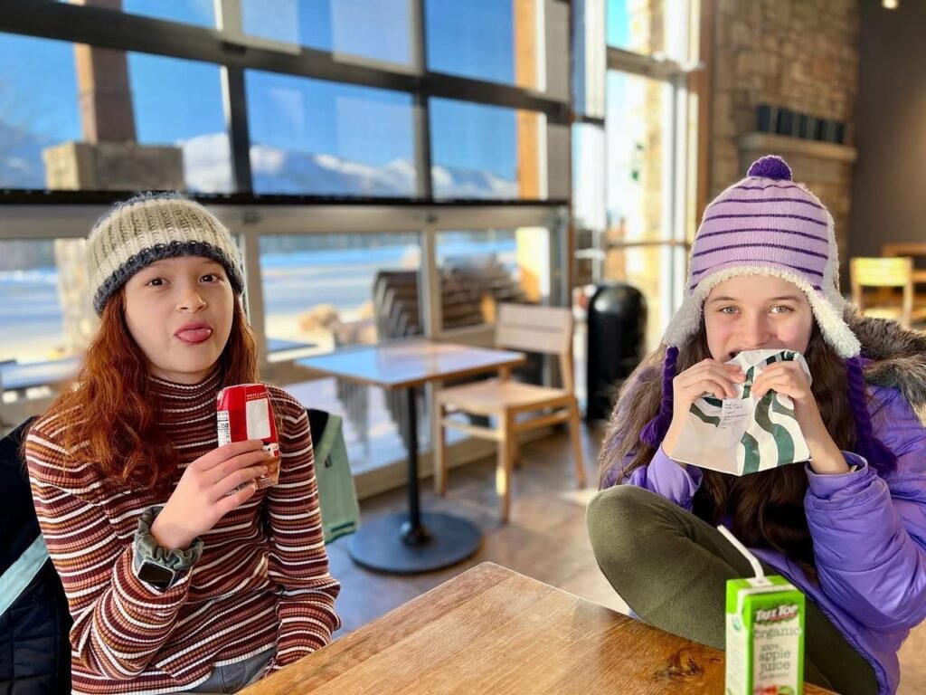 Two people enjoying a warm drink at a table in a light-filled cafe with large windows showing a mountain view.