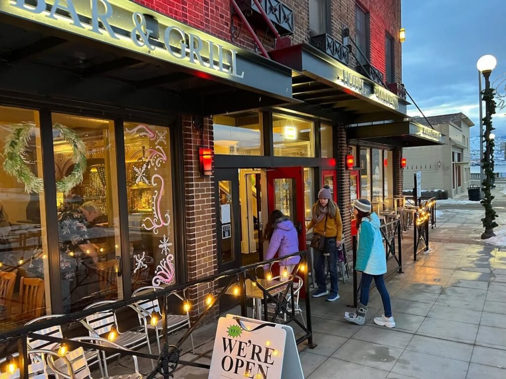 A street view of Park & Chisel restaurant with outdoor seating and a wet, reflective sidewalk. Warm string lights illuminate the chairs and tables at dusk, creating a welcoming atmosphere with holiday decorations visible in the windows.