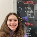 A young person with long hair smiles in front of a decorative name board with handwritten lists.