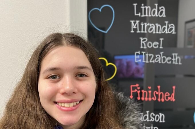 A young person with long hair smiles in front of a decorative name board with handwritten lists.