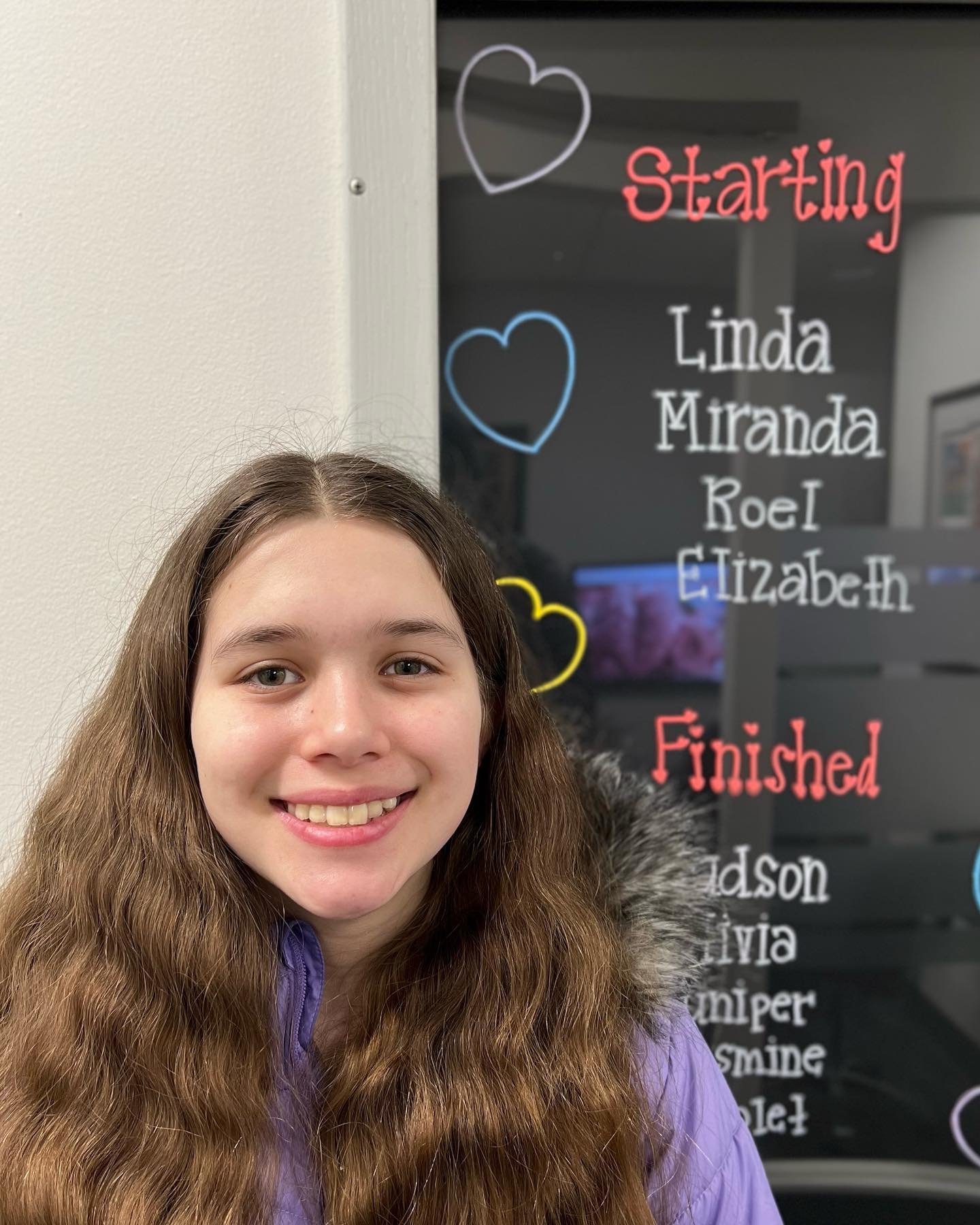 A young person with long hair smiles in front of a decorative name board with handwritten lists.