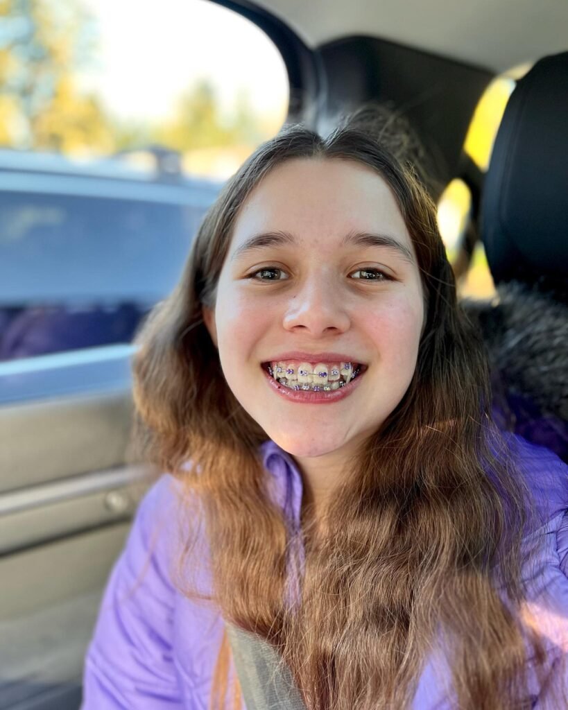 A smiling person wearing braces sits in the back of a car with autumn foliage visible outside.