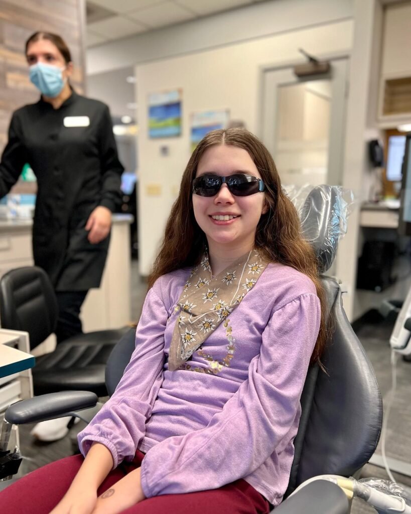 A young person wearing special eye protection smiles while seated in an optical clinic. A medical professional in the background wears a face mask during the visit.