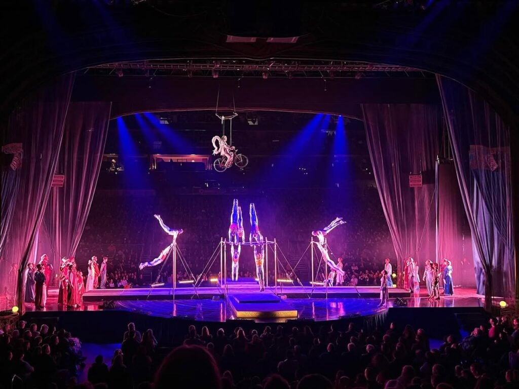 Acrobats perform high above a circus stage illuminated by vibrant blue and purple lighting while an audience watches from the arena floor.