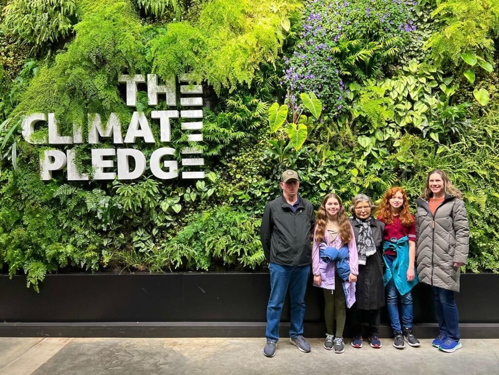 A group stands in front of the Amazon Climate Pledge logo on a living wall. The lush greenery provides a vibrant backdrop for the photo.