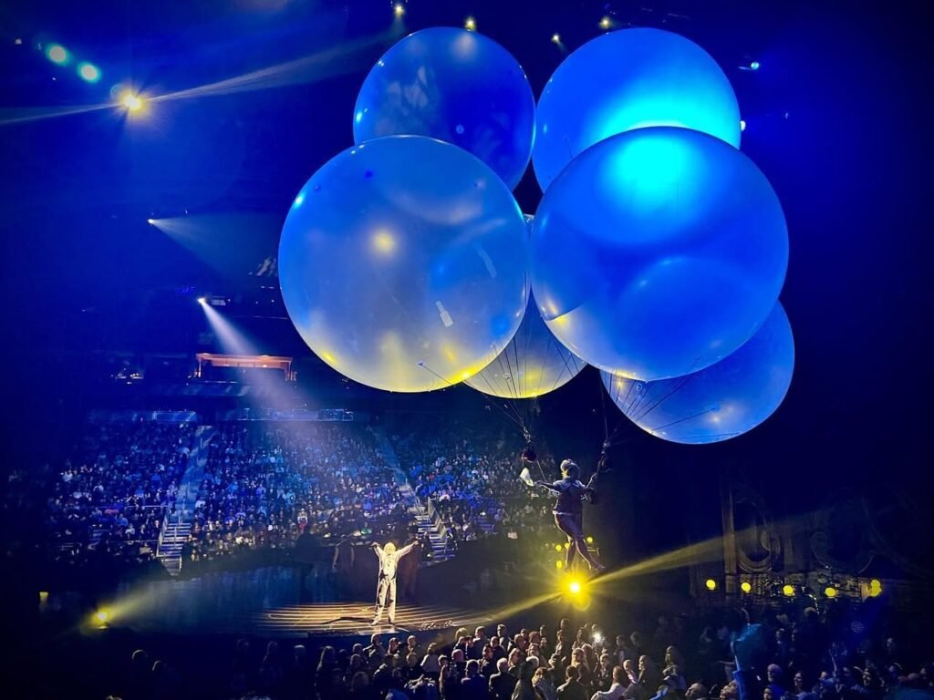 A dramatic stage performance featuring large blue balloons and bright lighting over an audience.
