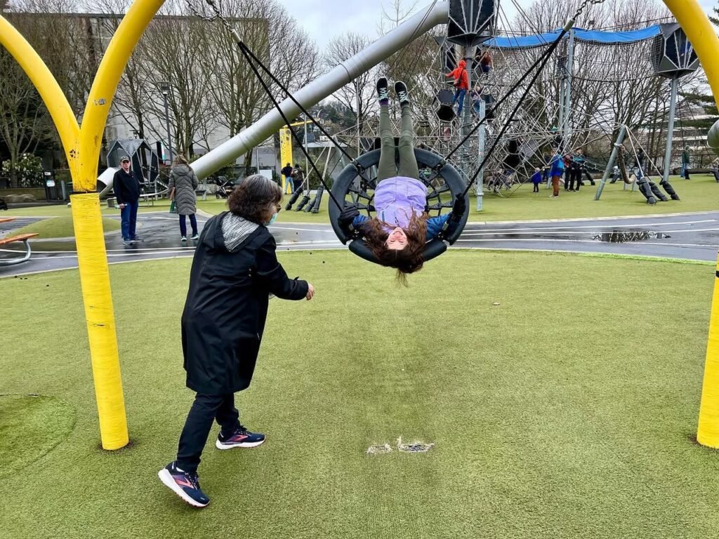 A child upside down in a spinning swing at SplashPlay while another plays on the turf. The playground features yellow poles and large equipment.