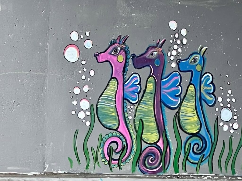 A vibrant mural featuring three seahorses in pink, purple, and blue with sea grass and bubbles. A whimsical example of urban street art on a grey wall.