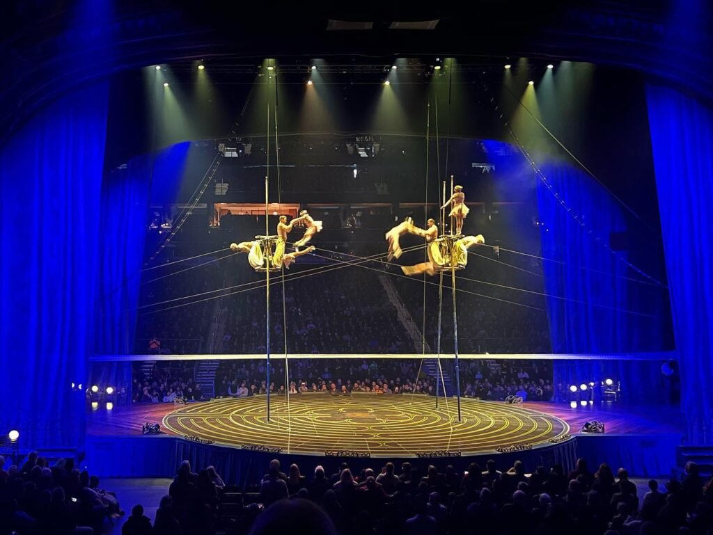 Aerial acrobats perform above the grand ring under vibrant blue lighting.