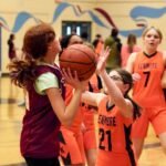 San Juan Island Spring Break A young basketball player in a maroon jersey gently embraces a teammate from the Linmore team, who wears an orange jersey with the number 21.
