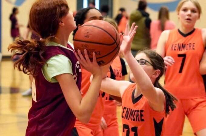 A young basketball player in a maroon jersey gently embraces a teammate from the Linmore team, who wears an orange jersey with the number 21.