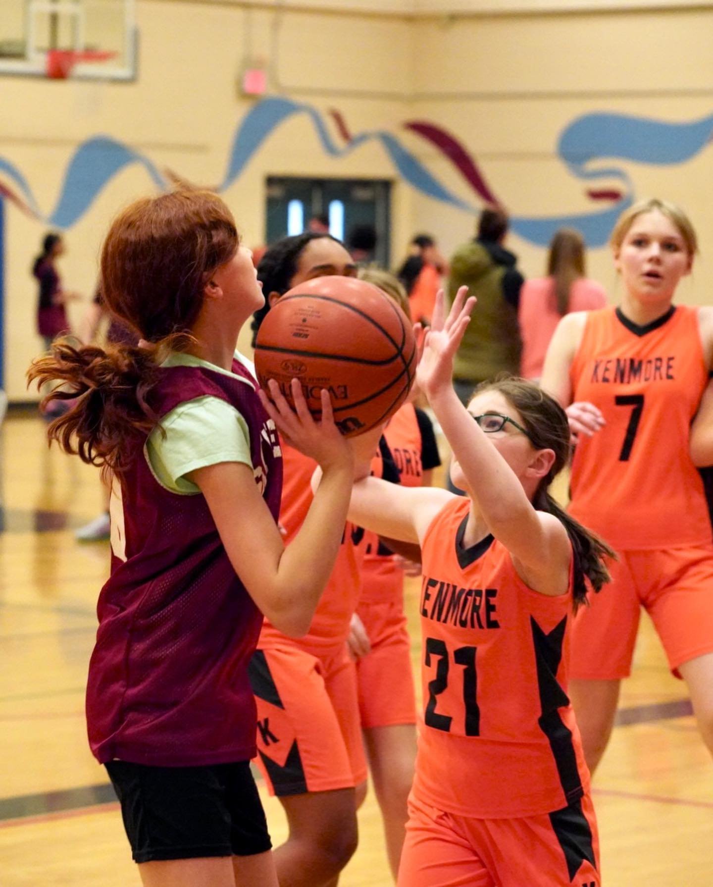 A young basketball player in a maroon jersey gently embraces a teammate from the Linmore team, who wears an orange jersey with the number 21.