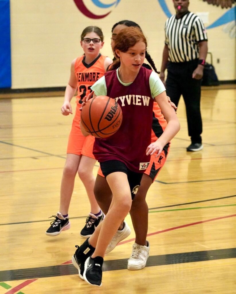 Player in a maroon jersey dribbles a basketball while being guarded by a player in an orange jersey during an indoor game.