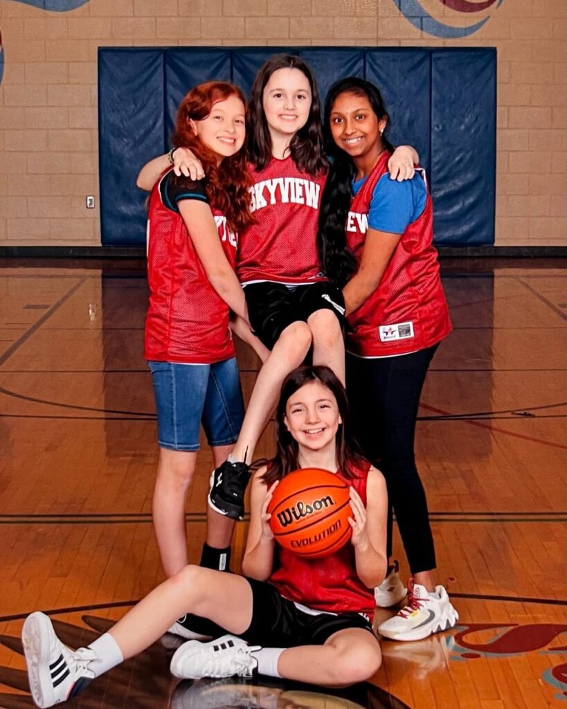 Group of young basketball players posing with an orange Wilson basketball in a gymnasium. They are wearing red jerseys and athletic gear on a hardwood court.