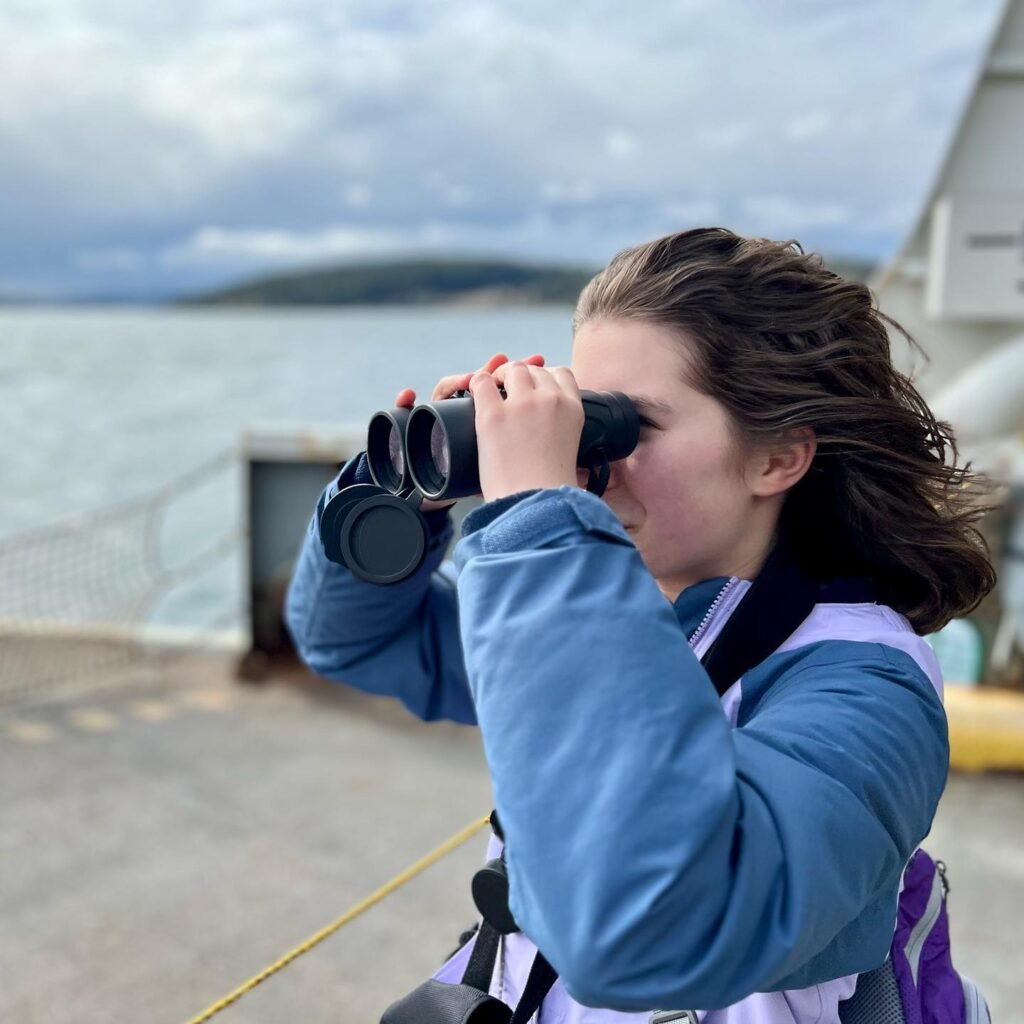 A person uses binoculars to view a misty coastal landscape in the Pacific Northwest.