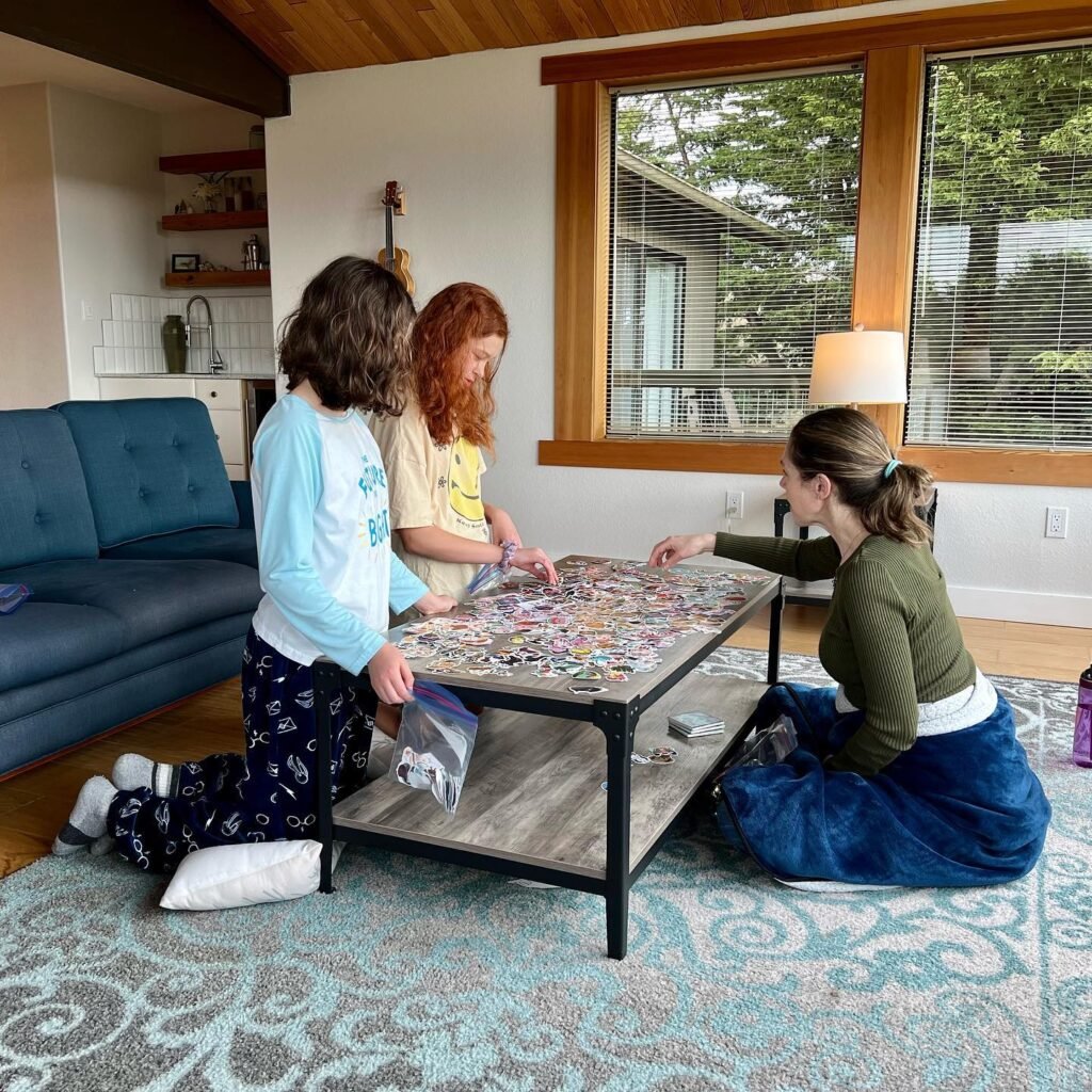 Three people work on a puzzle at a low table while sitting on the floor in a bright living room.