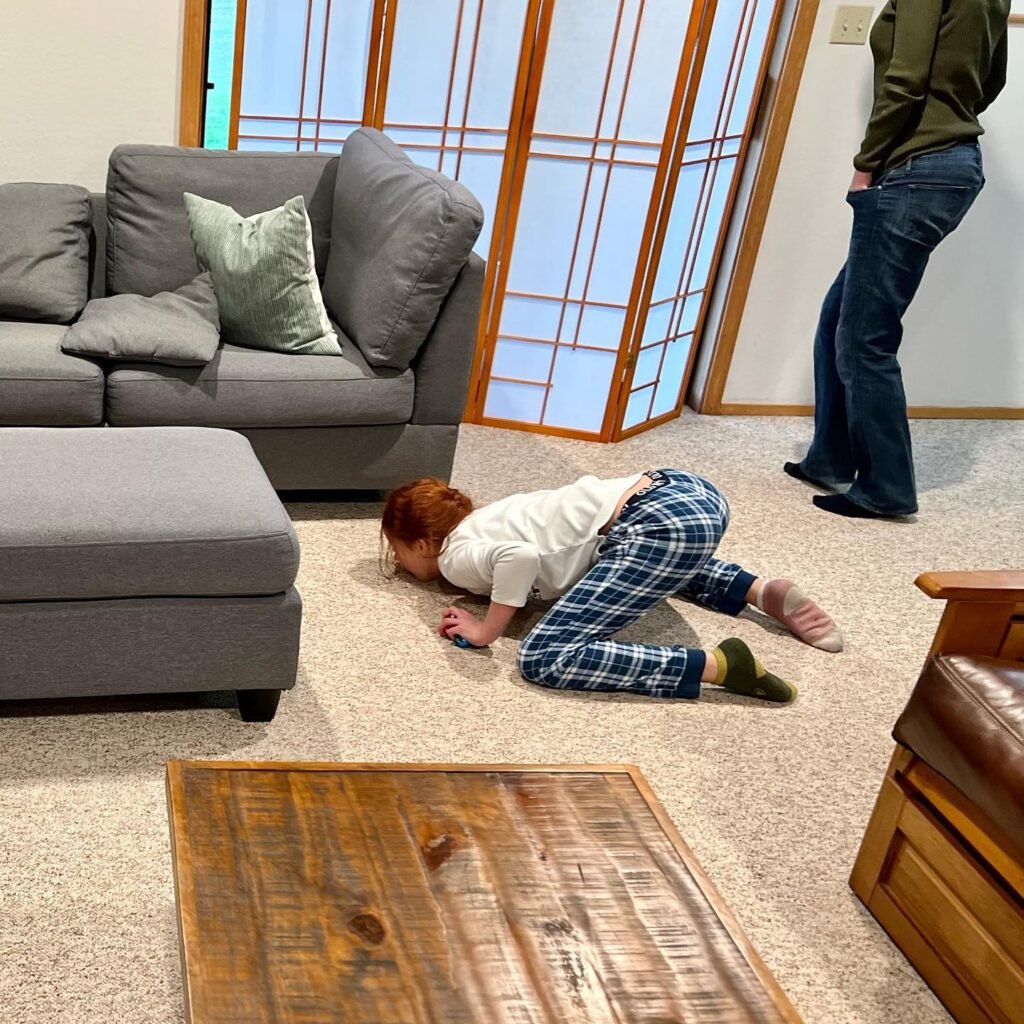 A young child crawls on carpet in a living room with gray couch and wooden table.