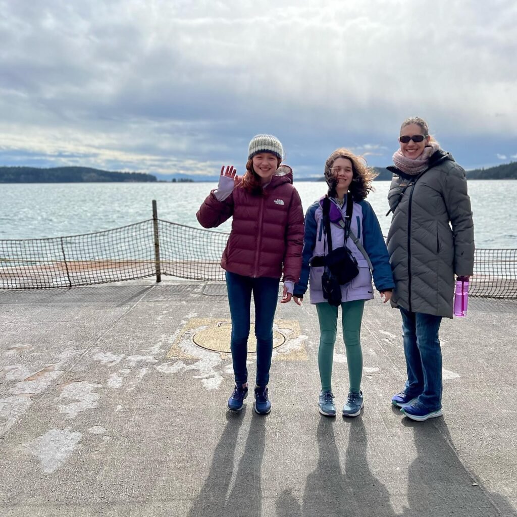 Three people in winter clothing pose and wave on a concrete pier with a large body of water and distant hills in the background under a cloudy sky.