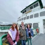 Three travelers in winter coats stand smiling on the deck of a white and green ferry against a cloudy sky. A child holds a camera while the family enjoys their boat trip to explore the waterways.