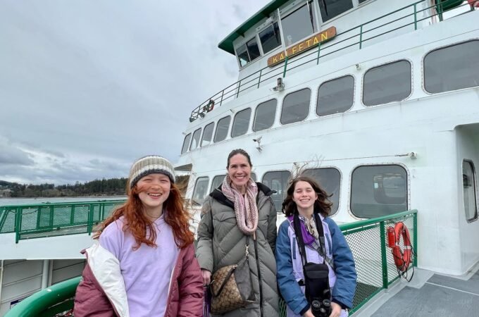 Three travelers in winter coats stand smiling on the deck of a white and green ferry against a cloudy sky. A child holds a camera while the family enjoys their boat trip to explore the waterways.