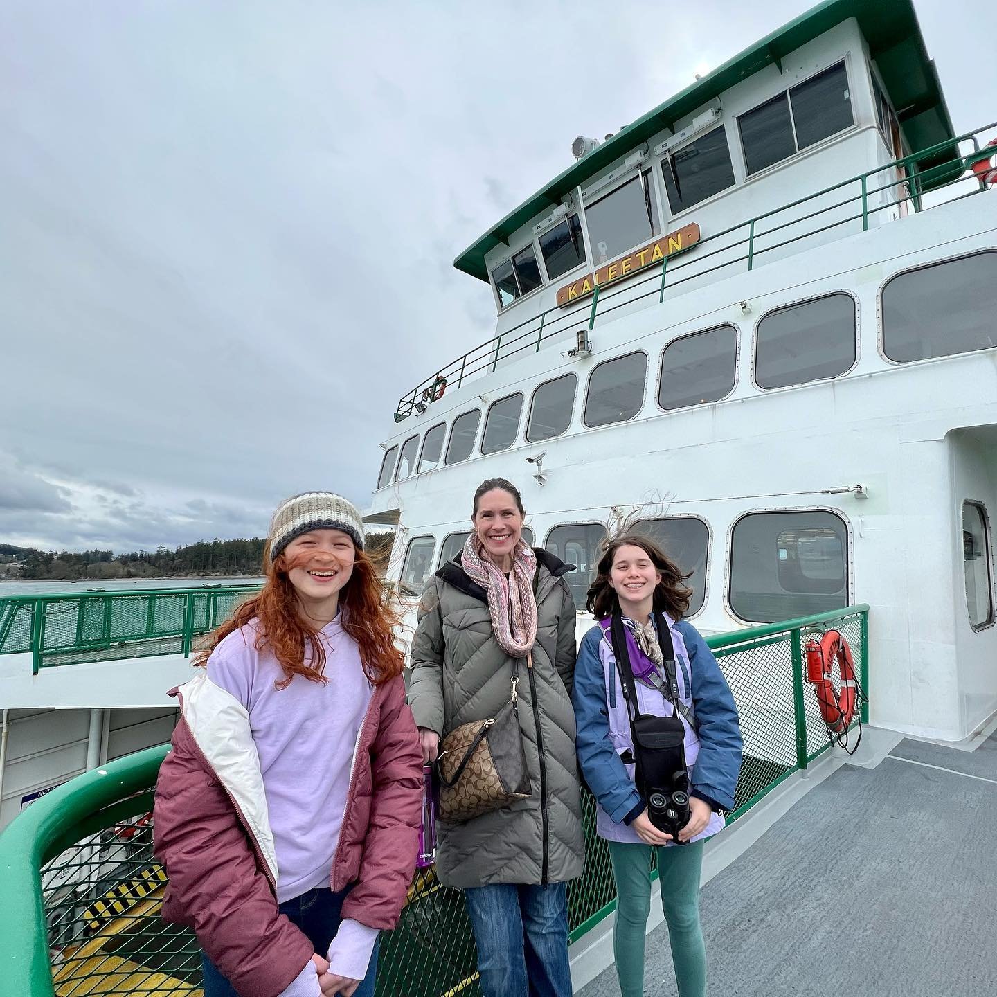 San Juan Island Spring Break Three travelers in winter coats stand smiling on the deck of a white and green ferry against a cloudy sky. A child holds a camera while the family enjoys their boat trip to explore the waterways.