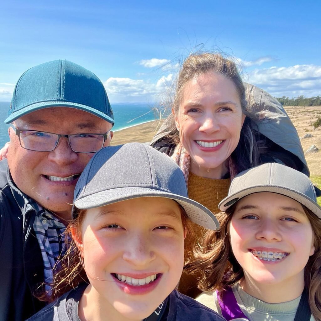 A smiling family takes a selfie together against a backdrop of the Pacific Ocean and rolling coastal hills. Bright blue skies and golden light complete the scenic view.