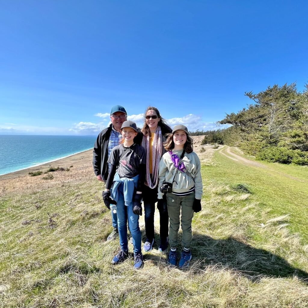 A smiling family poses together in a grassy coastal area overlooking a calm, deep blue ocean under a clear sky.
