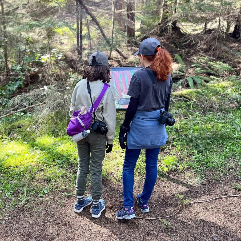 Two hikers in outdoor gear with cameras and packs stand on a forest trail near an information panel in a lush forest setting.