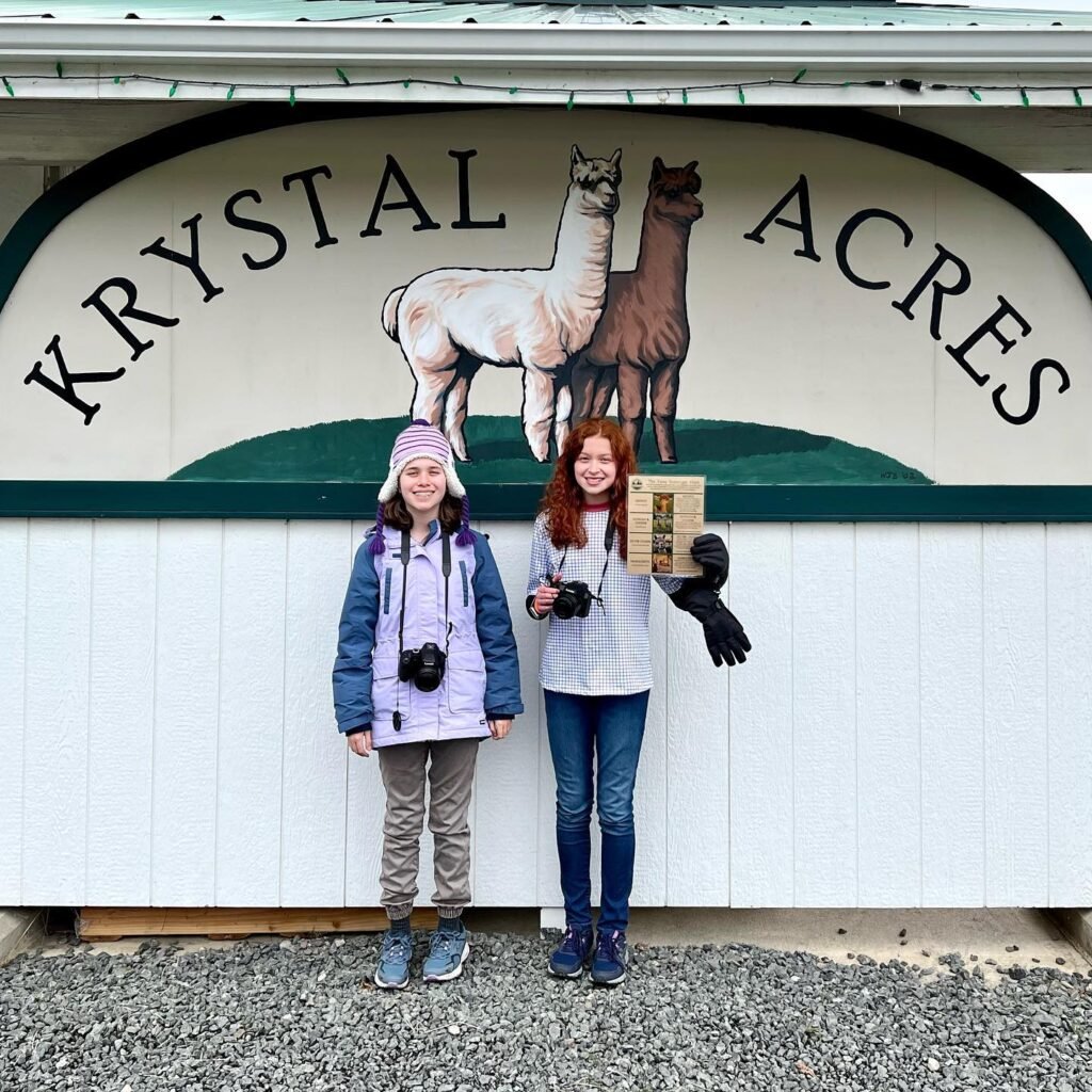 Two visitors standing in front of a building at Krystal Acres, with the business sign and alpaca illustrations visible above them in the Pacific Northwest.