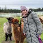 A visitor in winter gear smiles next to a wire fence while interacting with alpacas on a farm.