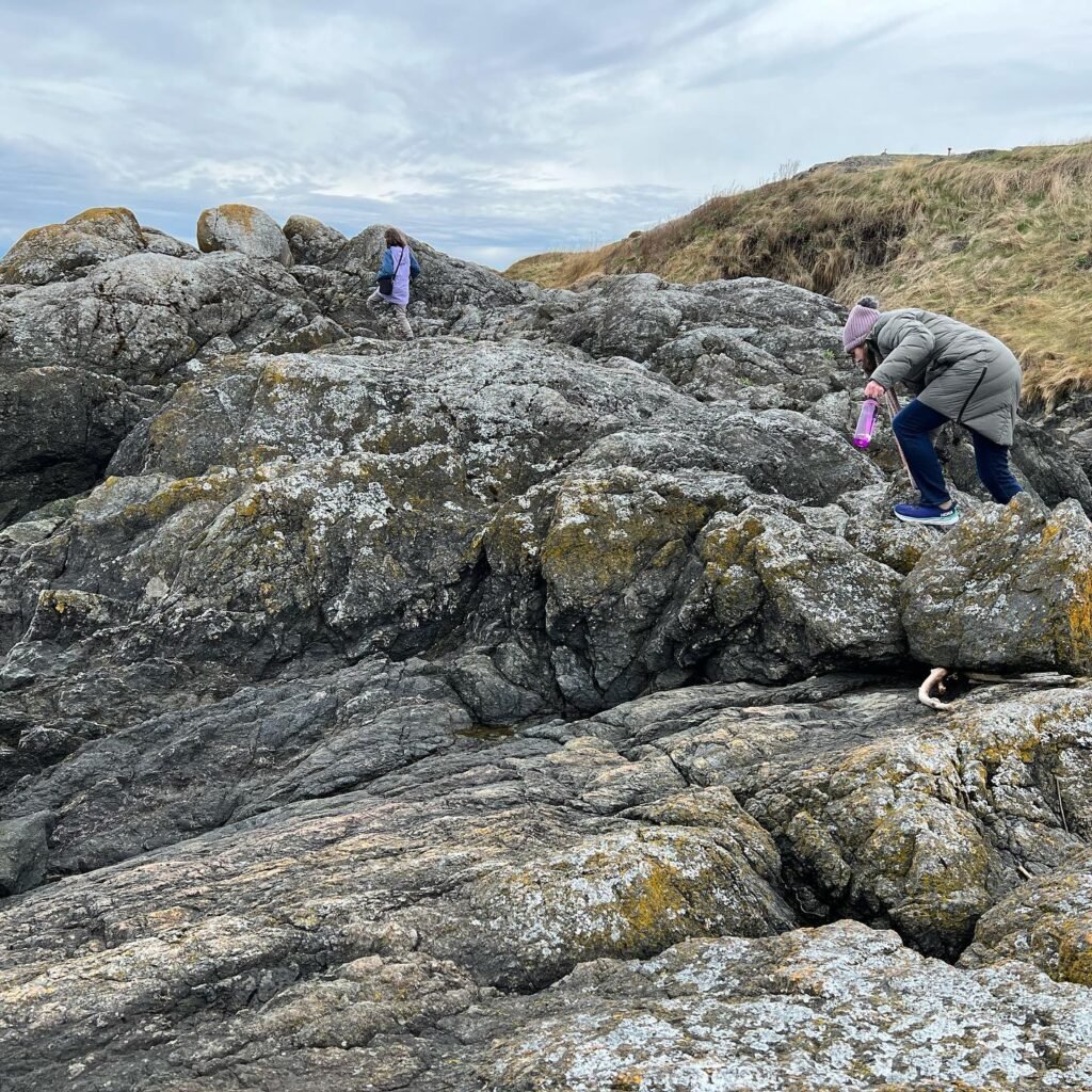 Travelers navigate rugged coastal rocks under an overcast sky. Durable footwear and patience are needed for this terrain.