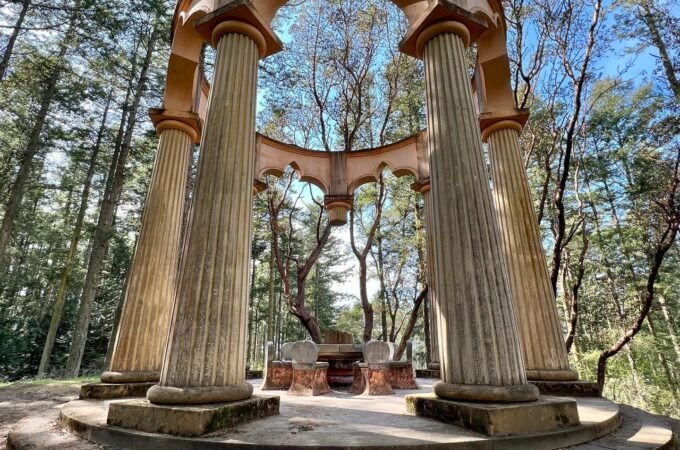 A panoramic view of a circular stone gazebo with fluted columns nestled in a forest. The structure features arched openings and steps leading up to a central platform.
