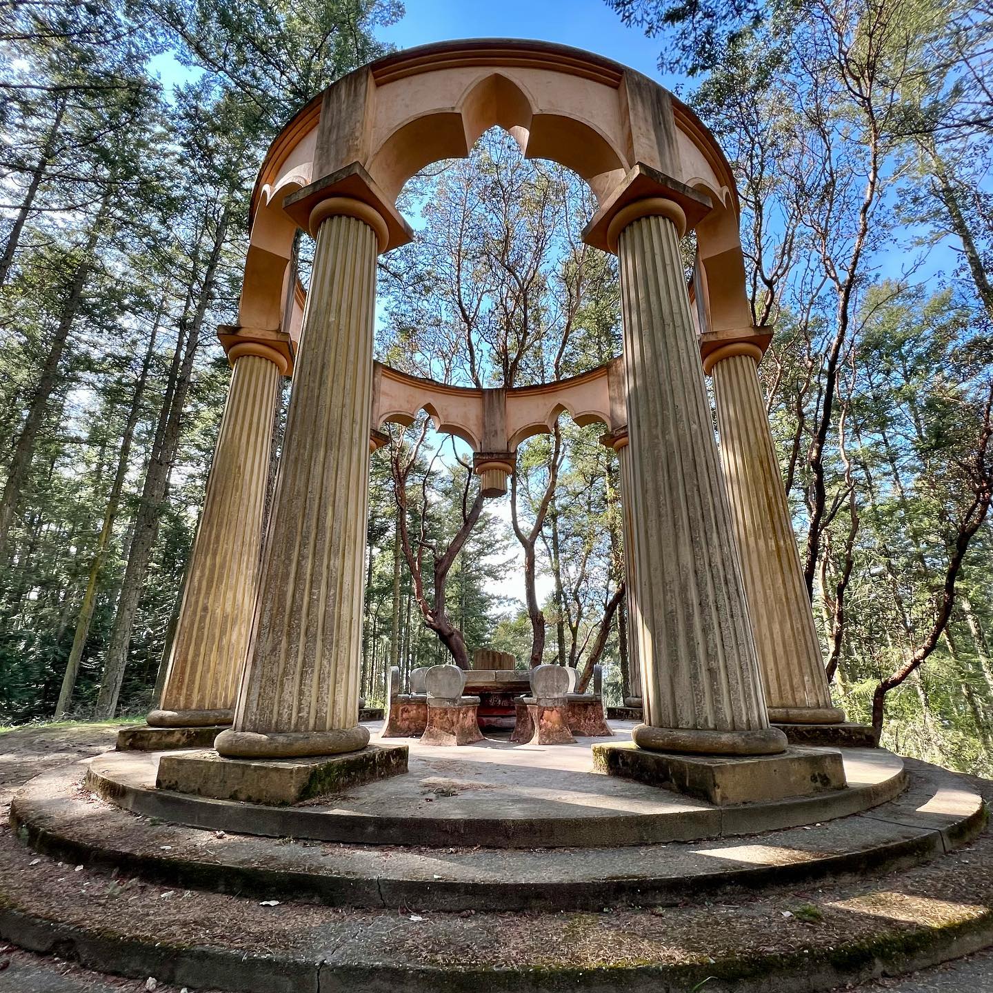 A panoramic view of a circular stone gazebo with fluted columns nestled in a forest. The structure features arched openings and steps leading up to a central platform.