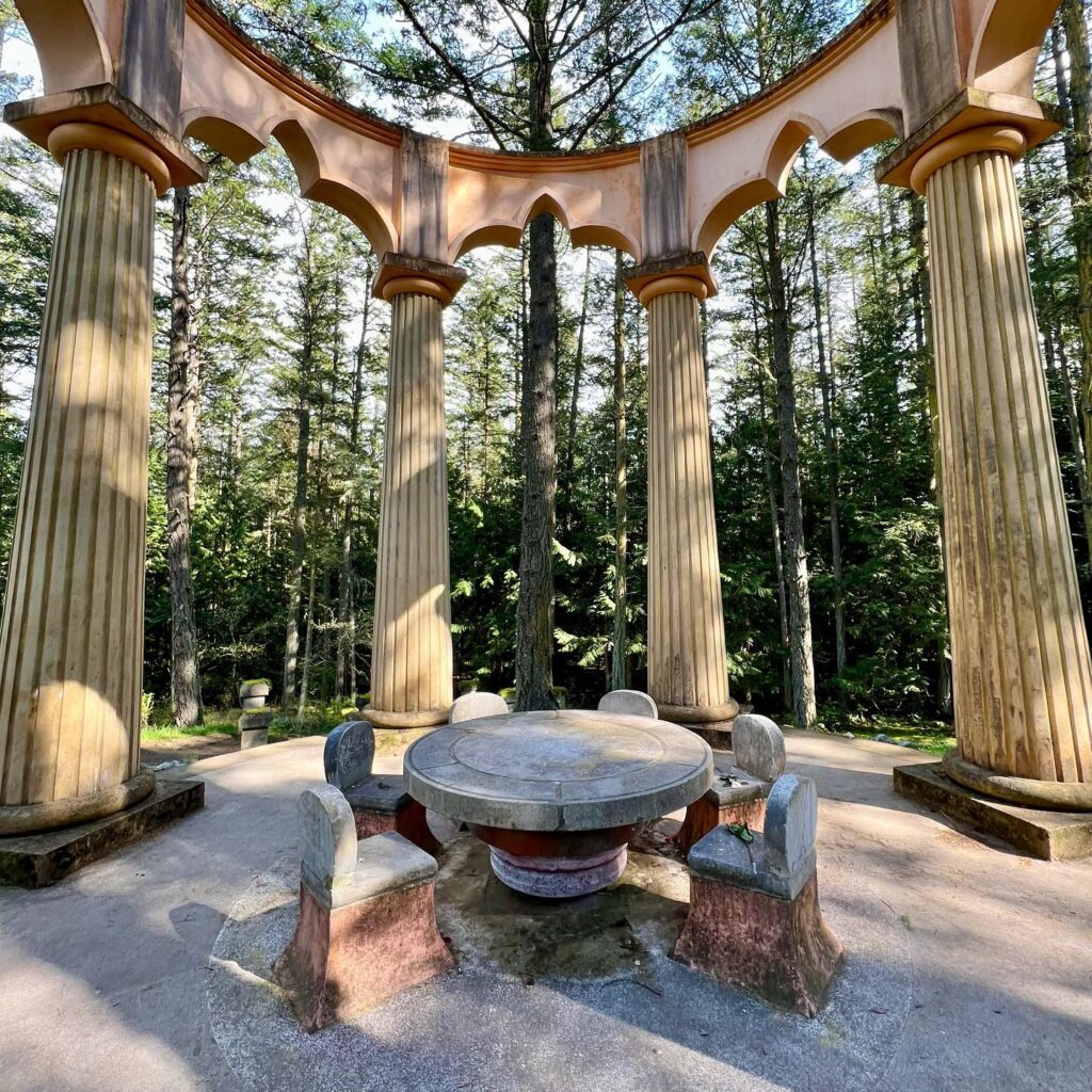 A detailed view of a rustic wooden gazebo featuring tall columns and an arched roof. The structure is surrounded by tall pine trees in a serene forest setting.